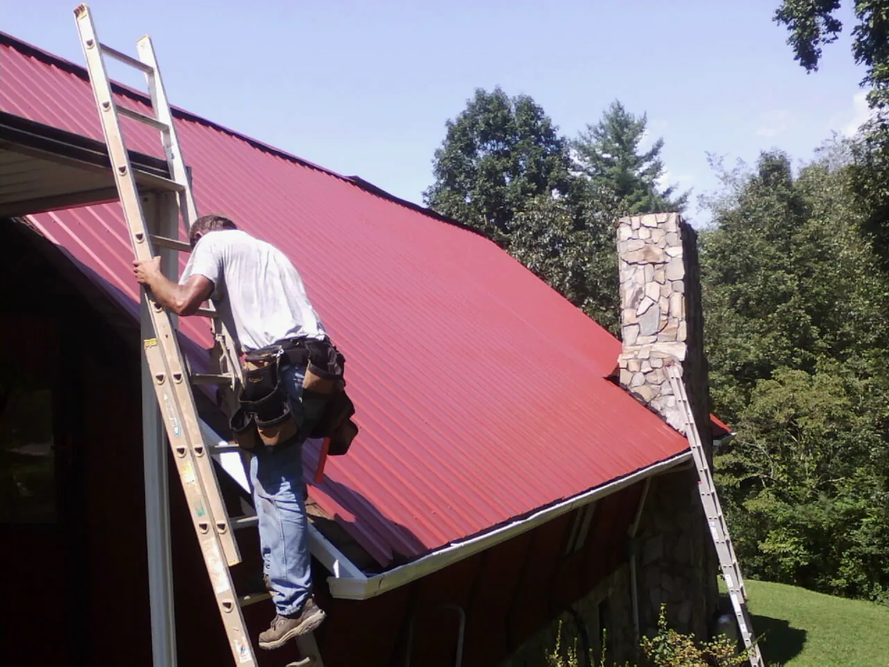 Roofer on a ladder beside a red metal roof and stone chimney — Soffit & Fascia Repair by Roofing Brothers in Jordan Valley